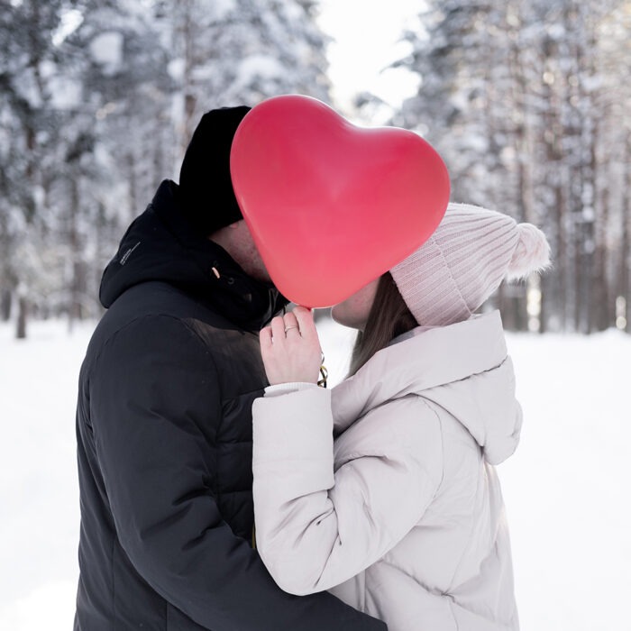 A couple holds a heart shaped balloon while standing in a snowy forest.